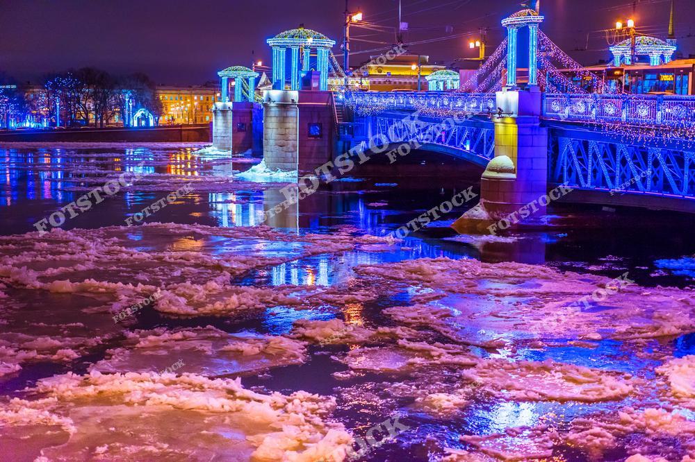 На фотографии показан мост в Санкт-Петербурге, украшенный праздничной иллюминацией в зимний вечер. Яркие огни синего и розового оттенков отражаются на поверхности реки, заполненной снежными льдинами. Иллюминация добавляет мосту особую сказочность, подчёркивая красоту архитектуры. Этот вид символизирует зимний Петербург, наполненный атмосферой праздника и уюта.